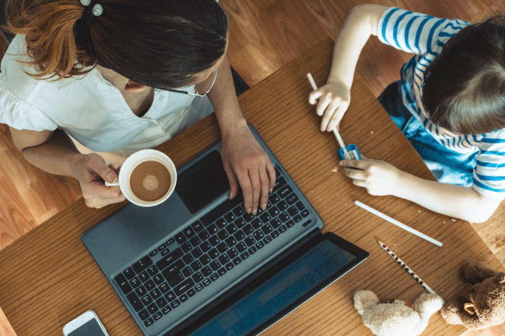 A mother works at a laptop with a cup of coffee while her young child draws at the same table
